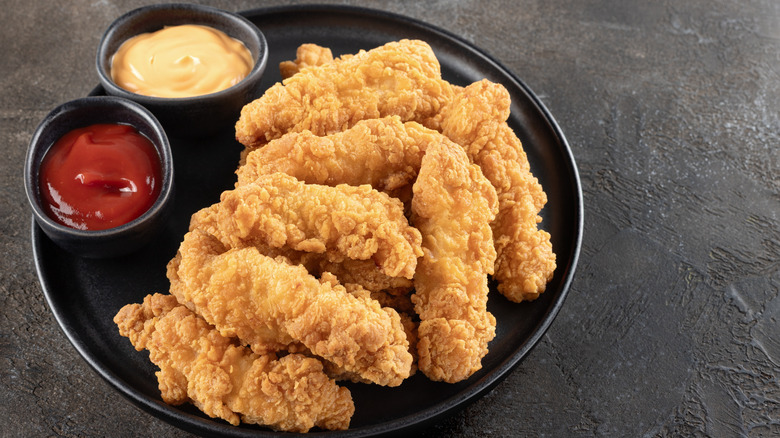Chicken fingers on a black ceramic plate with ramekins of ketchup and another side sauce
