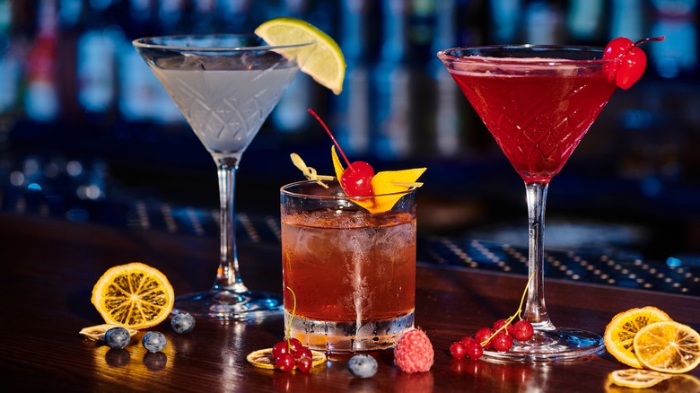 Three cocktails with fruit garnishes are lined up on a bar