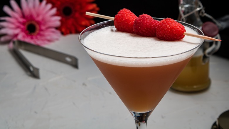 A pink, frothy cocktail in a martini glass garnished with three raspberries