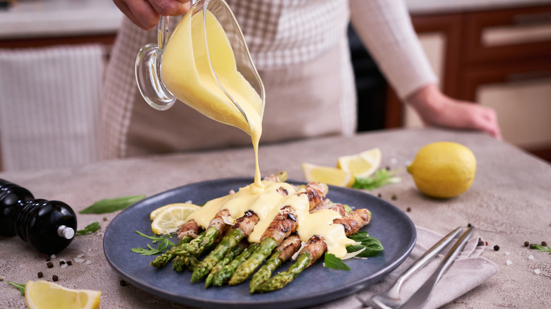 A cook pouring hollandaise sauce over a plate of asparagus stalks wrapped in bacon