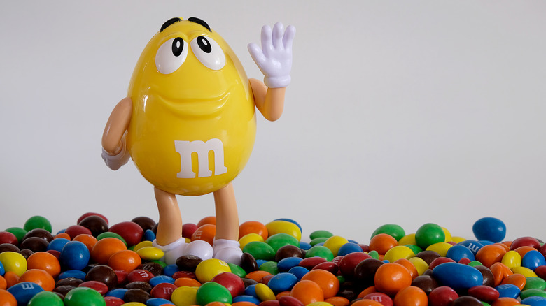 A figurine of the yellow peanut M&M standing on a pile of M&M's
