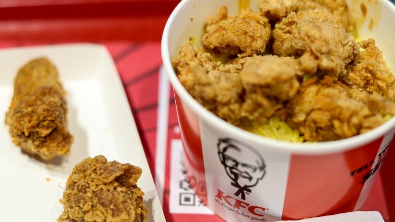 A bucket of KFC fried chicken on a red tray with two pieces out next to it.