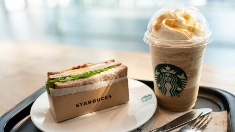 A sandwich from Starbucks on a plate sitting next to a frappucino on a tray with a set of silverware.