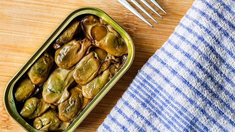 An open tin of canned oysters on a wooden table next to a napkin and fork.
