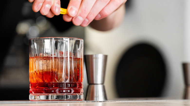 A person's hands are seen squeezing an orange peel over a cocktail in a rocks glass.