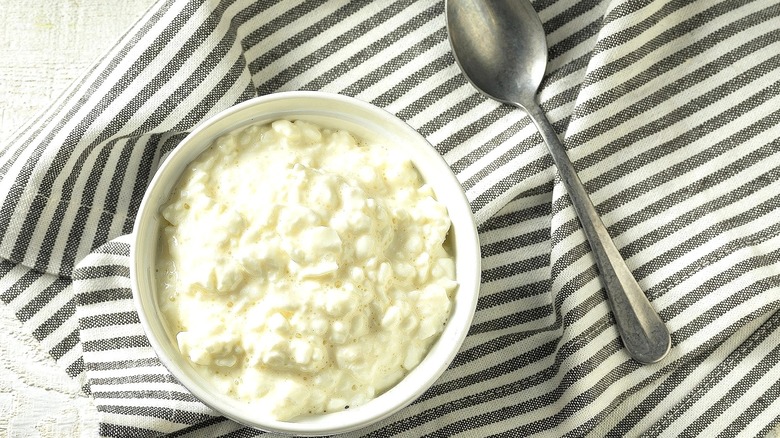 Cottage cheese in a white bowl next to a metal spoon over a gray-and-white-striped cloth napkin
