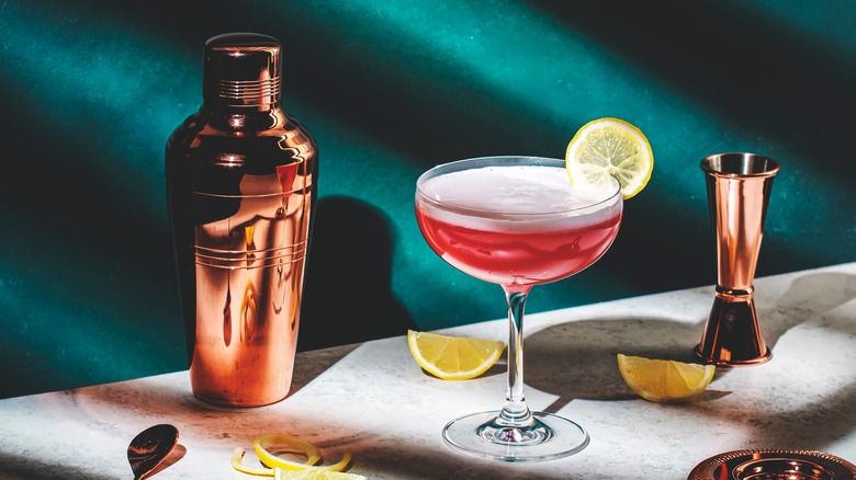 A copper cocktail shaker next to a frothy cocktail garnished with a lemon slice