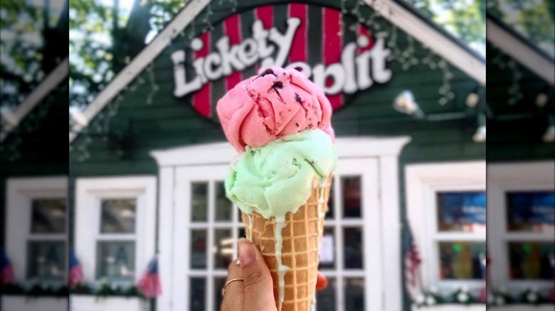 Person holding a double-scoop ice cream cone in front of Lickety Split ice cream, which looks like a tiny house behind a white picket fence.