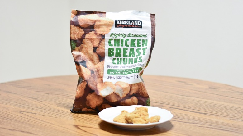 A bag of breaded chicken breast chunks behind a white plate of chicken pieces
