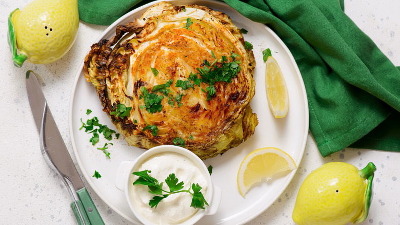 Cabbage steaks with a garnished pot of sour cream on a plate next to lemon wedges and a green cloth in the background