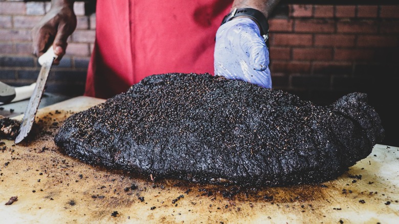 A cook holding up a whole, uncut smoked brisket on a cutting board