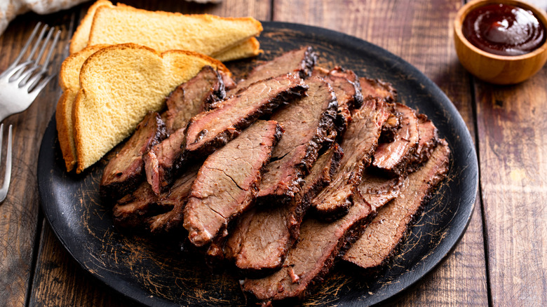 A black plate loaded with slices of brisket and Texas toast with BBQ dipping sauce in the background