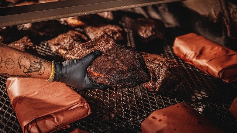 A pitmaster picks an unwrapped brisket up from a pit full of wrapped briskets