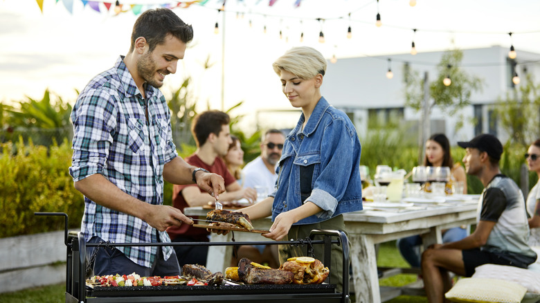 Two people serve themselves from a grill; more people sit at a picnic table behing them.