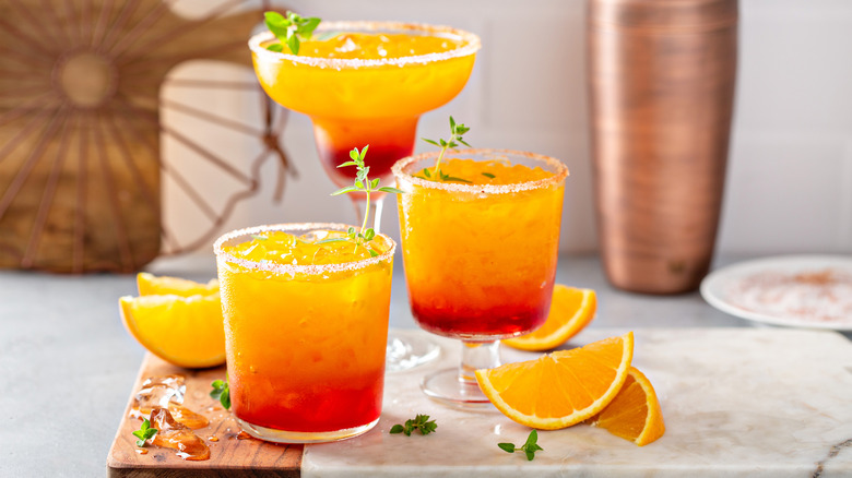 Tequila sunrise in different glasses surrounded by orange wedges placed on cutting board