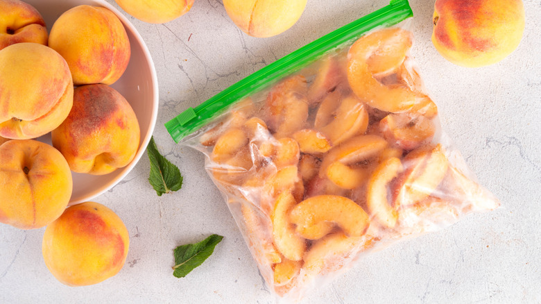 Zippered bag filled with frozen peach slices next to several whole, fresh peaches