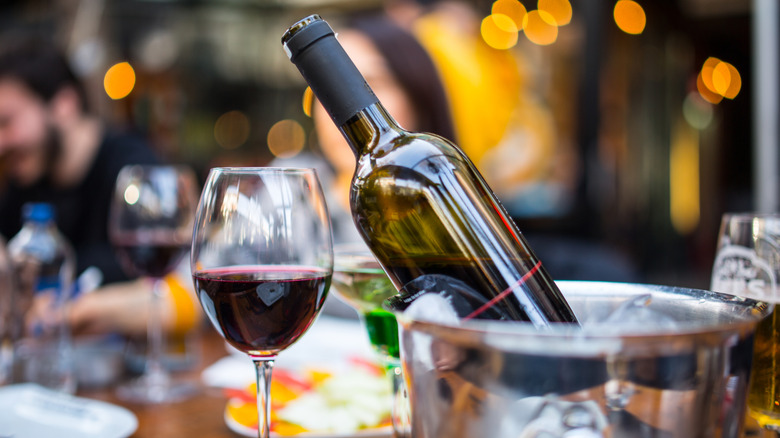 A glass of red wine is beside a bottle chilling in an ice bucket on a restaurant table.