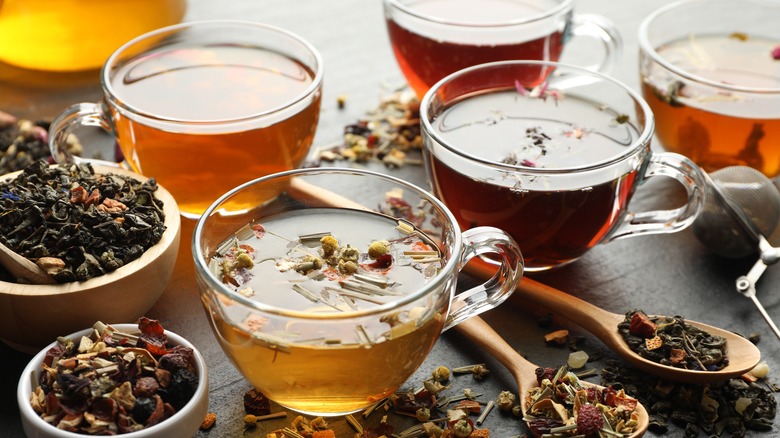 Several glass cups of tea surrounded by bowls and spoons filled with tea leaves and herbal tea