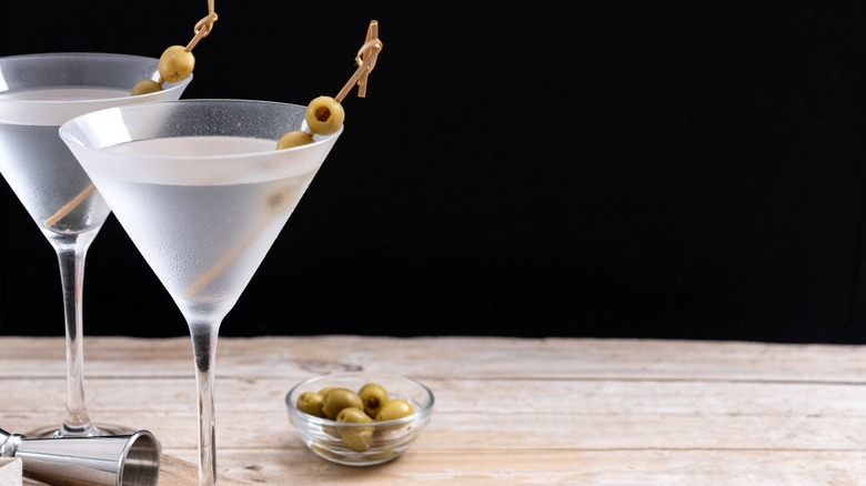 Two vodka martinis with olives on toothpicks and a bowl of olives on a wooden table