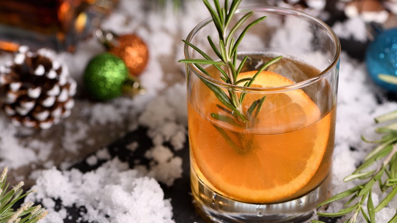 Glass of bourbon with an orange segment and rosemary sprig as garnishes in front of a Christmas backdrop with fake snow and ornaments