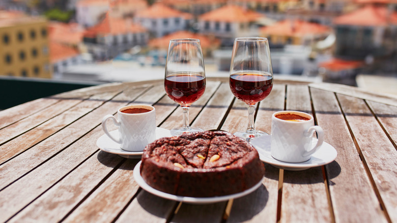 Two glasses of Madeira wine with two white cups of espresso on white saucers and a plate with a Portuguese honey-nut cake on a table outdoors