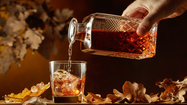 Hand pouring whiskey from decanter to glass on a wood table next to dry leaves