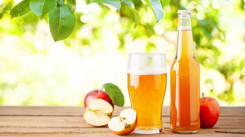 A bottle and glass both filled with hard cider on a table next to two whole apples and two halves, under a leafy tree on a sunny day