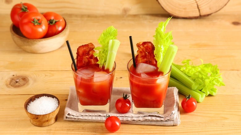 Two Bloody Mary drinks, each with a stirrer and celery stick, next to a bowl of cherry tomatoes, more celery, and salt