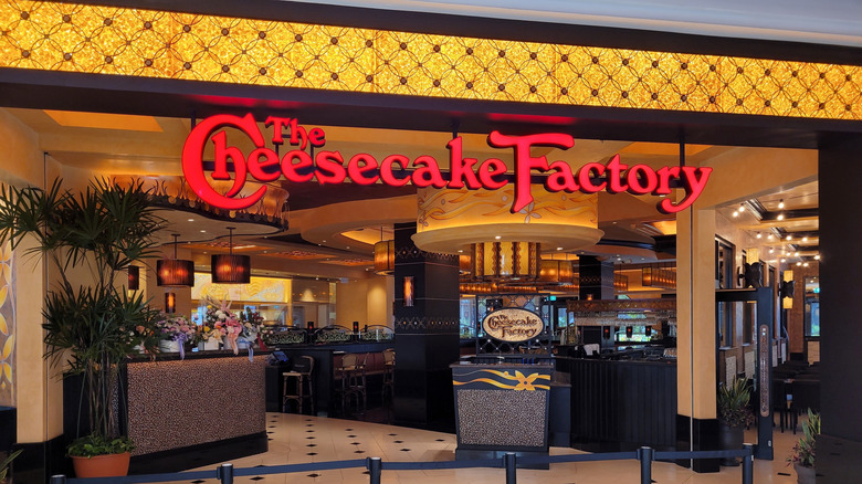 A Cheesecake Factory restaurant interior as seen from its entrance, with a lit Cheesecake Factory sign above it.