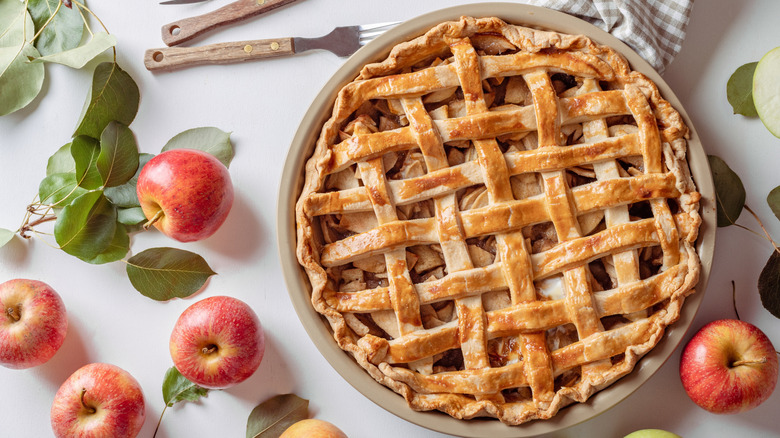 Apple pie with lattice crust surrounded by apples, utensils, and leaves.