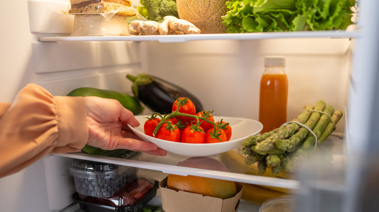Person's hand reaching into fridge full of produce for a bowl of tomatoes