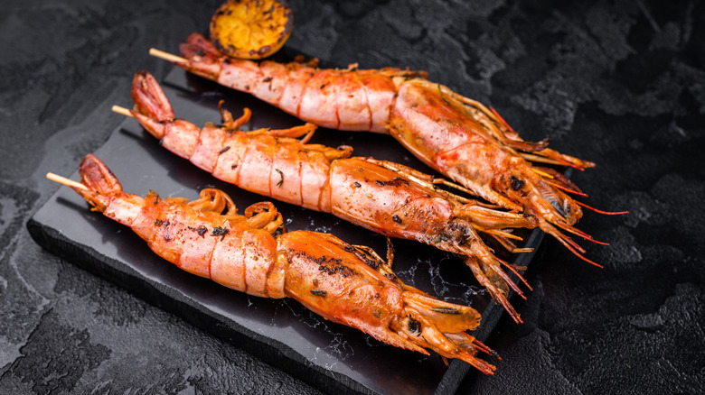 Three red langostinos are skewered and arranged on a black stone board with a charred lemon half.