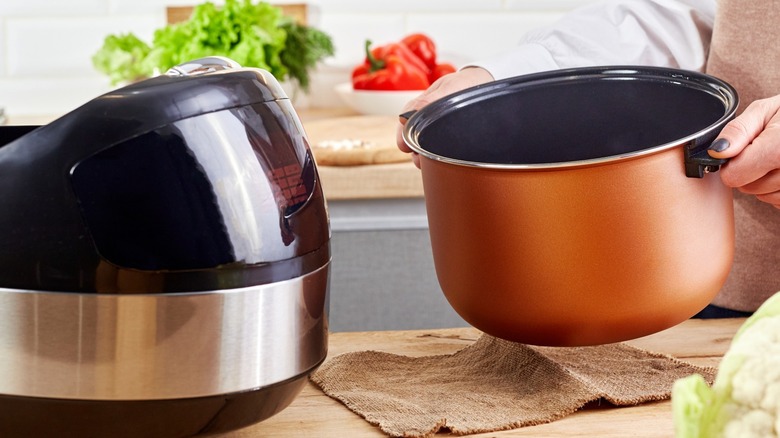 Woman placing ceramic pot into slow cooker on wooden countertop.