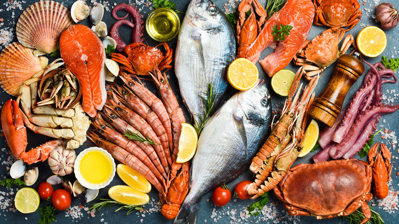 A variety of fresh, raw seafood displayed on a table with scattered ingredients like salt, herbs, tomatoes, and lemons
