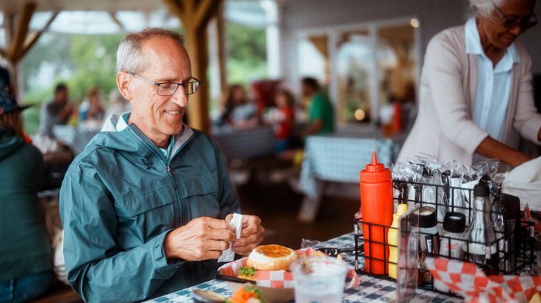 An elderly man eating at an American restaurant outside with people in the background.