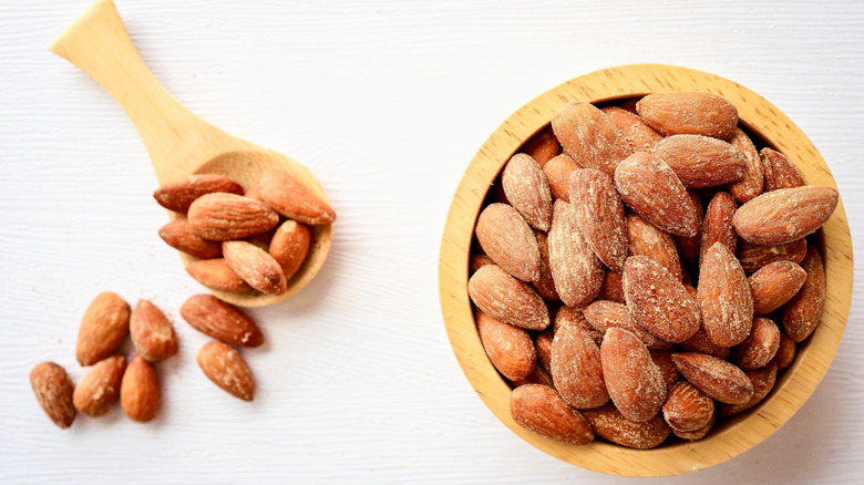 Salted, roasted almonds in a wooden scoop and a wooden bowl on a white surface