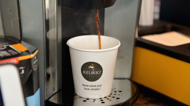 A Keurig coffee maker pouring hot coffee into a paper cup with its logo.