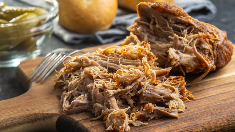 Delicious, perfectly cooked smoked pulled pork on a cutting board, pickles and buns behind it.