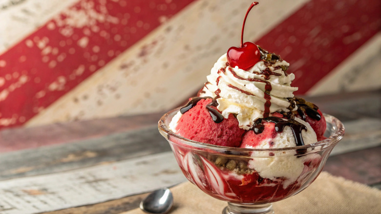 A classic ice cream sundae in a glass dish with strawberry and vanilla ice cream, topped with whipped cream, chocolate sauce, and a cherry on top, served on a rustic wooden table with a striped wooden panel behind.