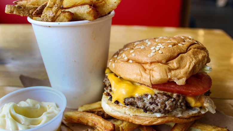 A cheeseburger with lettuce and tomato on a sesame seed bun sits beside a cup of hand-cut french fries and a smaller cup, which contains a spread that looks like mayonnaise, on a restaurant table. More fries lie underneath the burger.