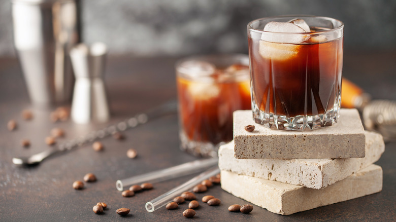Iced coffee liqueur on counter next to coffee beans with shaker tin in background.