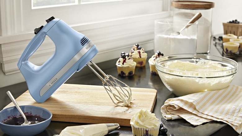 5-Speed KitchenAid hand mixer in blue velvet resting on a cutting board with a bowl of frosting and cupcakes around it