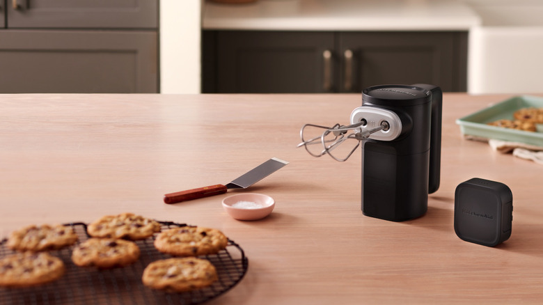 Black cordless hand mixer standing on end next to a cooling rack of chocolate chip cookies