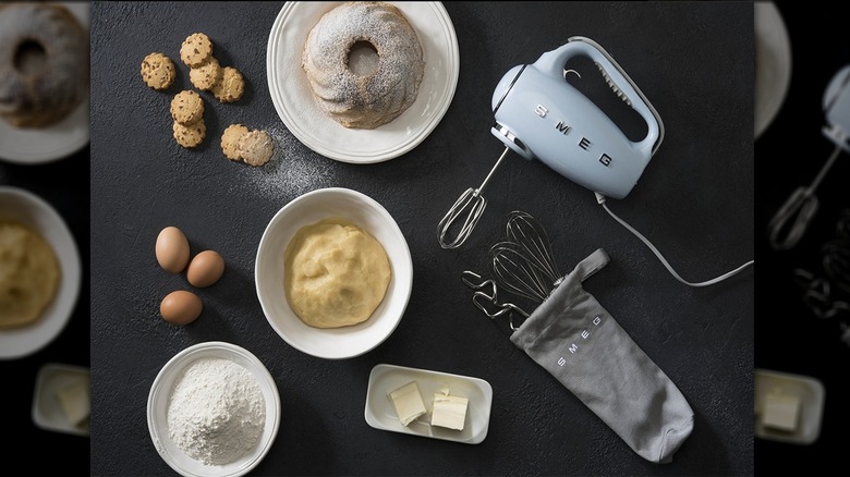 Pastel blue SMEG hand mixer lying on surface with bag of accessories, bowls of ingredients, and treats