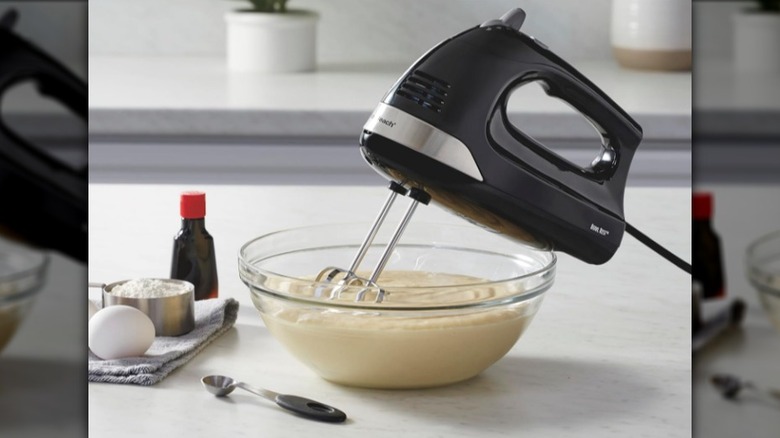 Black Hamilton Beach 6-Speed Hand Mixer with beaters in a glass bowl full of light yellow batter next to an egg, vanilla, and measuring cup of white powder