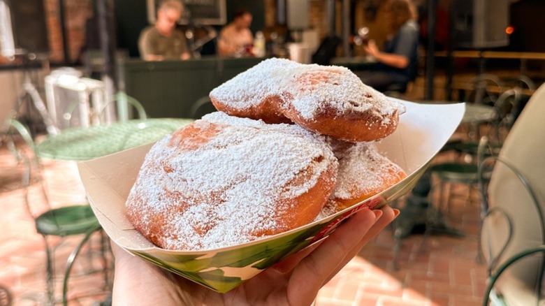 A paper plate with three sugar-dusted beignets being held up with restaurant tables in the background
