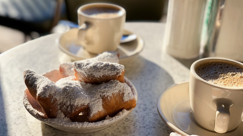 Round table with white plate holding 5 beignets, plus two white cups/sauces containing cafe au lait, sugar and napkin containers in the background