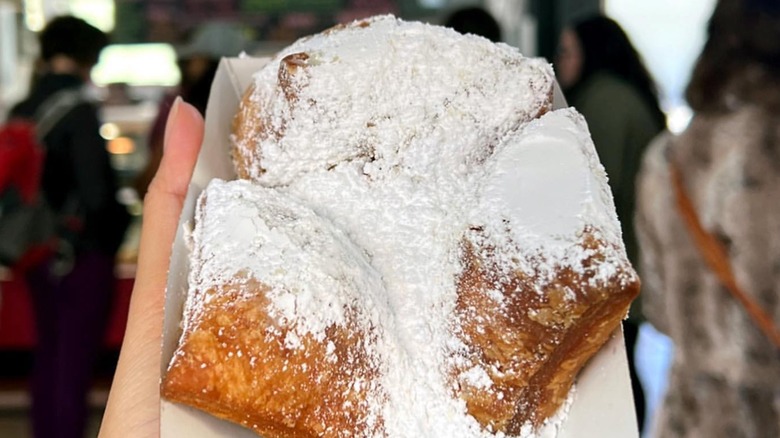 Three sugared beignets in a paper tray being held up by hand with restaurant in background