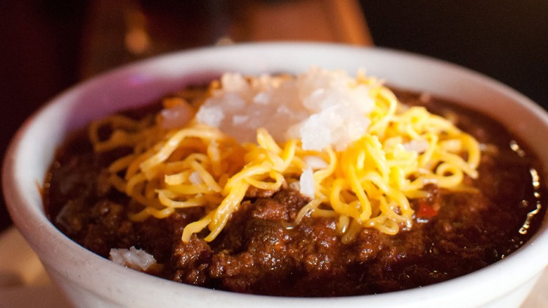 White bowl of chili topped with shredded cheese and diced white onion