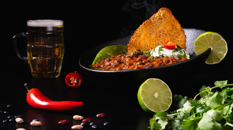Bowl of chili on black background with limes, peppers, and a pint of beer
